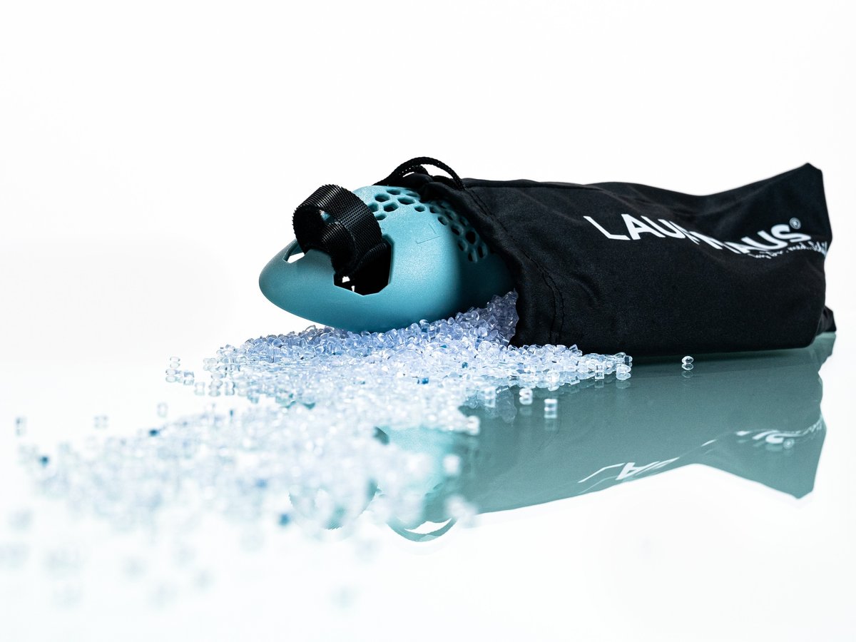 Une souris de course bleue, de forme ergonomique, dépasse d'un sac en tissu noir portant l'inscription blanche « LAUFMAUS ». De petits granulés de plastique transparents sont dispersés sur une surface réfléchissante, sur fond blanc.