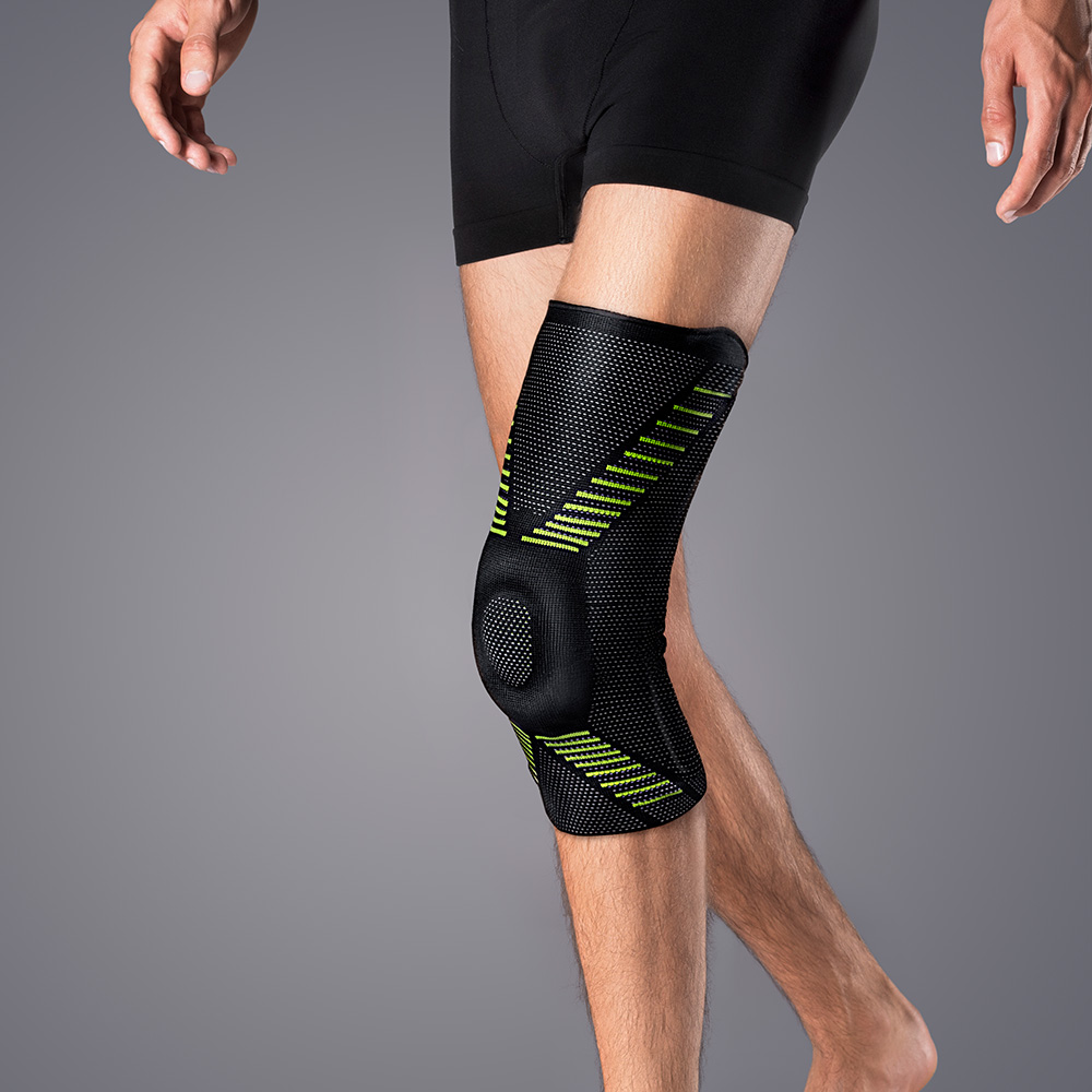 A leg from the hip to the foot on a grey background. The person is wearing a black knee brace with bright green accent stripes around the knee. It fits snugly and has a round, slightly raised pad over the kneecap.