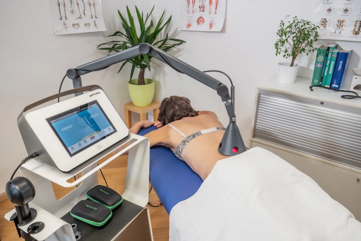 Person liegt bäuchlings auf blauer Behandlungsliege und erhält Lasertherapie am unteren Rücken. Grauer, schwenkbarer Laserarm mit rotem Strahl ist positioniert. Daneben modernes Gerät mit Touchscreen. Hintergrund: Wandbilder, Pflanzen.
