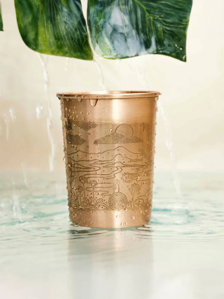 Eleganter Paradies-Kupferbecher mit handgeätzter Naturansicht aus Wolken, Bergen und Wasser. Der 300-ml-Becher ist mit Tautropfen bedeckt, steht in flachem Wasser, während von grünen Blättern Wasser herabrinnt.