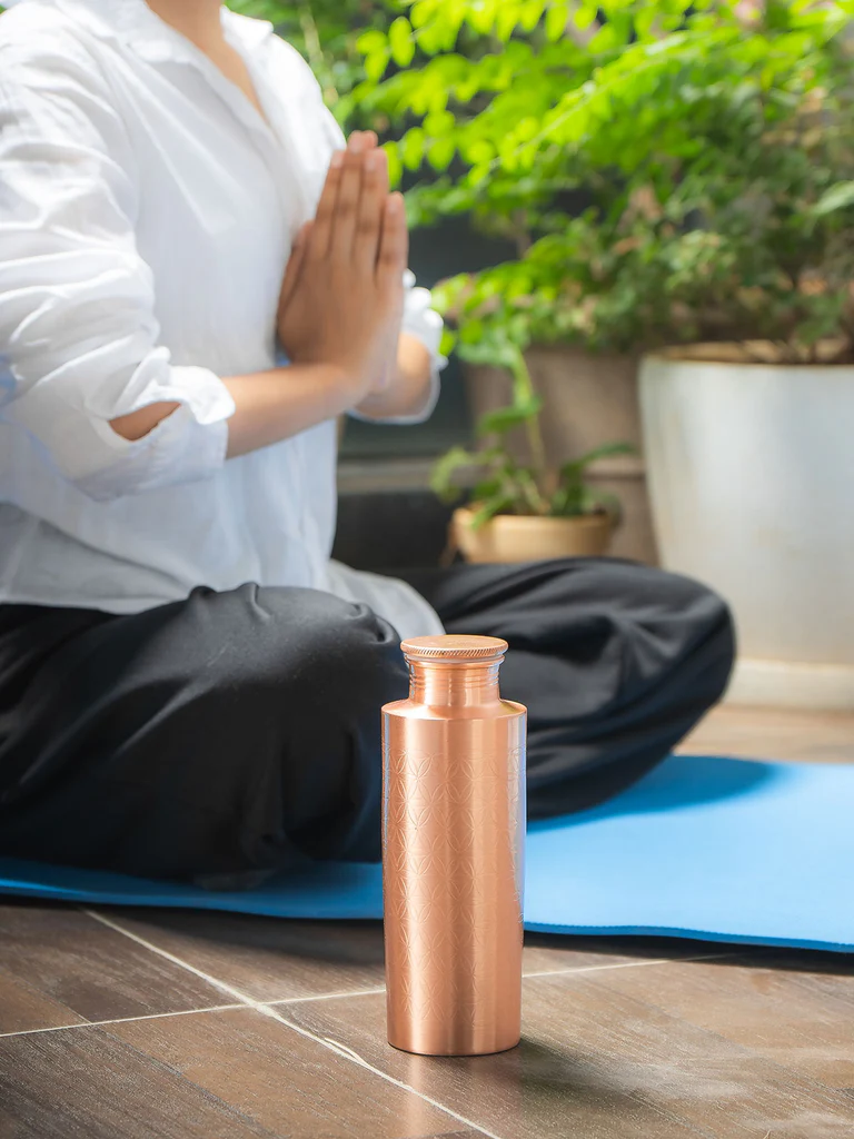 Glänzende Kupferflasche mit geprägtem Blume-des-Lebens-Muster steht auf Holzboden neben einer Person im Lotussitz auf blauer Yogamatte, Hände in Gebetshaltung vor der Brust. Im unscharfen Hintergrund: grüne Topfpflanzen.