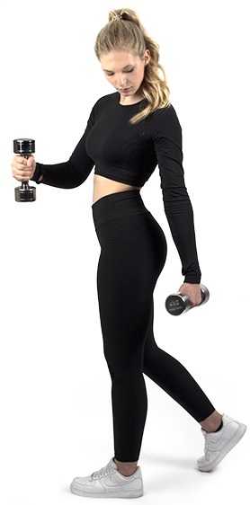 Jeune femme blonde en queue de cheval, tenue de sport noire (t-shirt manches longues ventre visible, leggings). Elle tient deux haltères chromés brillants à poignée striée et regarde celui de gauche. Baskets blanches.