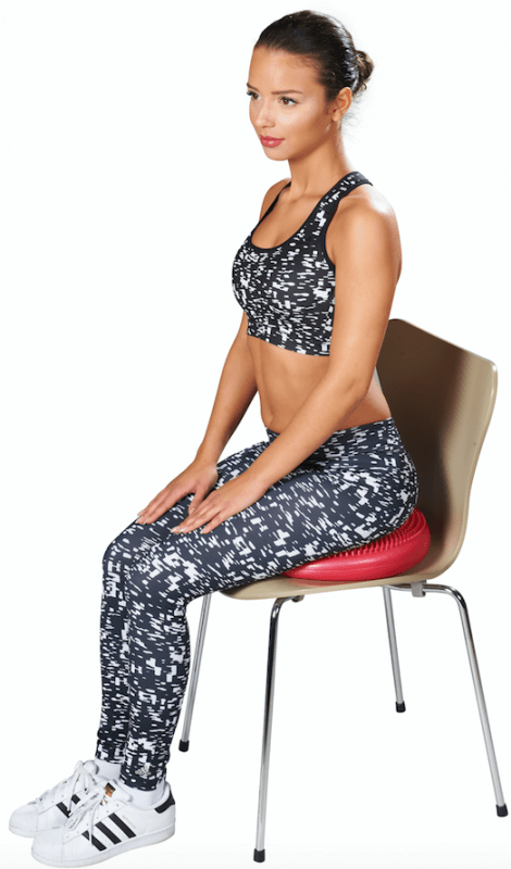 A young woman in black and white sportswear is sitting sideways on a brown chair and looking to the right. Beneath her on the seat is a red, round balance cushion with nubs, designed for dynamic sitting. Her hands are resting on her thighs.