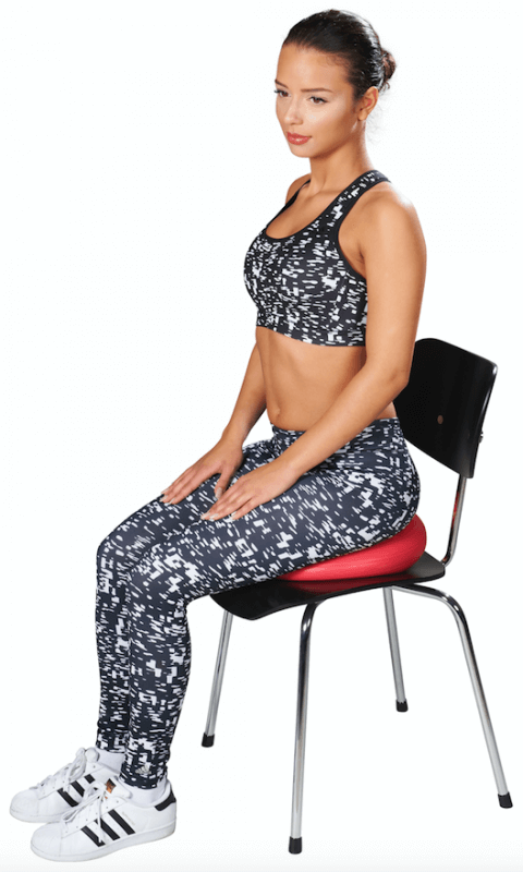 A woman in sportswear is sitting on a red balance cushion placed on a black chair, with her hands on her knees. The cushion promotes dynamic sitting, back stability and better posture.