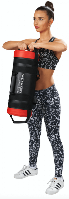 A young woman with her hair tied up, wearing sportswear, is holding a black cylindrical weight bag with red ends and the words ‘TRENDY ESFERA BAG’ printed on it in front of her with both hands; she is looking to the right against a white background.
