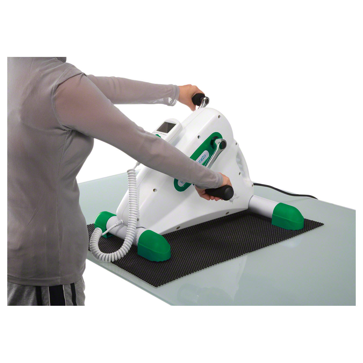 Eine Person trainiert ihre Arme mit dem weiss-grünen Arm- und Beintrainer Oxy Cycle auf einem Tisch, auf schwarzer rutschfester Matte. Sie hält die schwarzen Griffe, macht Rotationsbewegungen. Display und Kabel-Handschalter sichtbar.
