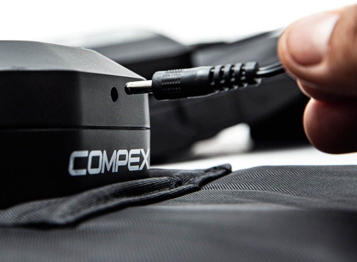 A finger is plugging a black charging cable into the charging socket of a dark Compex Ayre compression boot controller. The device, featuring a white Compex logo, is resting on black boot fabric and is being charged for wireless use.