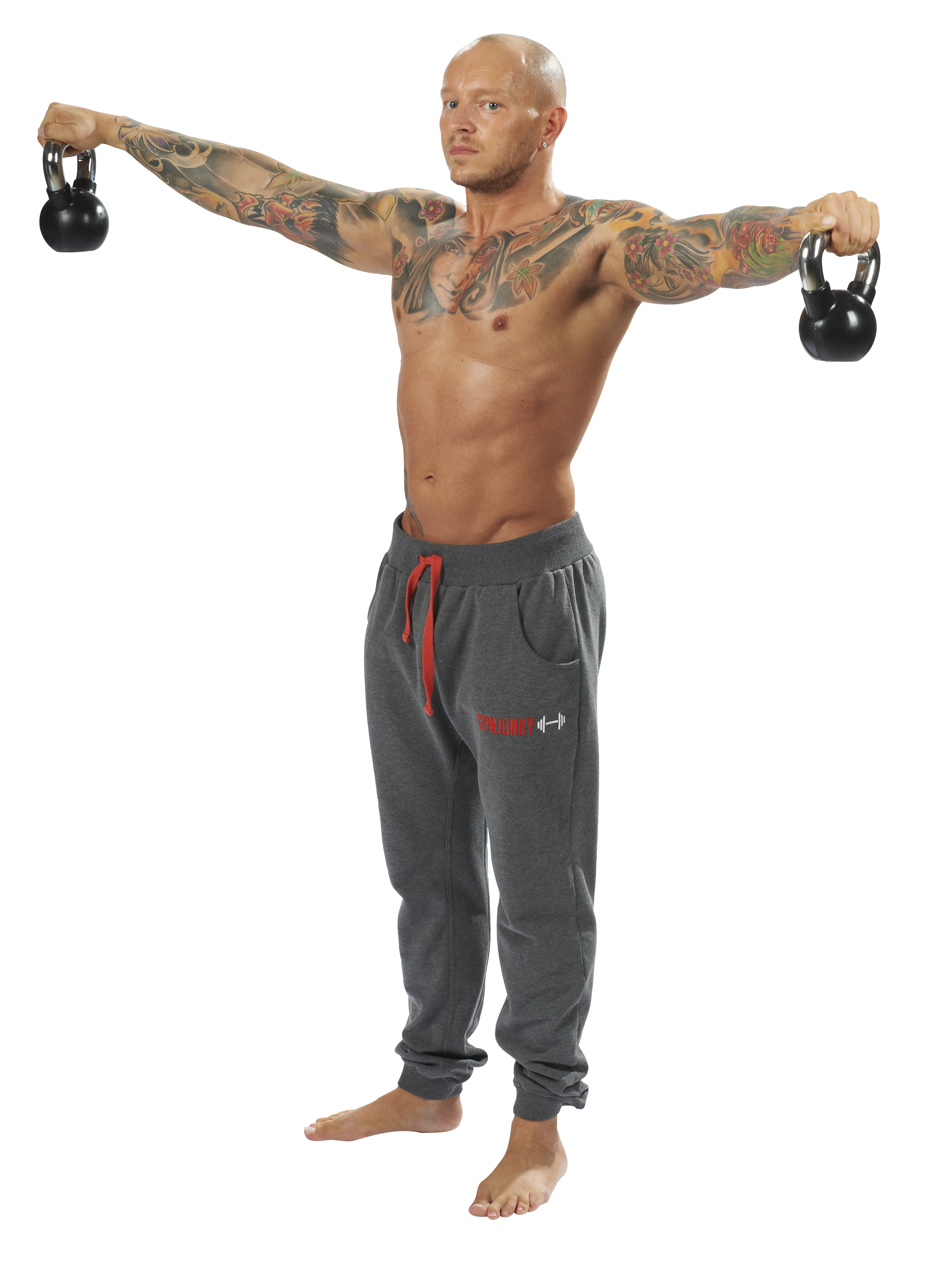 Un homme musclé et tatoué se tient pieds nus, les bras tendus, tenant un kettlebell noir dans chaque main. Torse nu, entièrement tatoué, il porte un pantalon de sport gris. Idéal pour l'entraînement de force et de coordination.