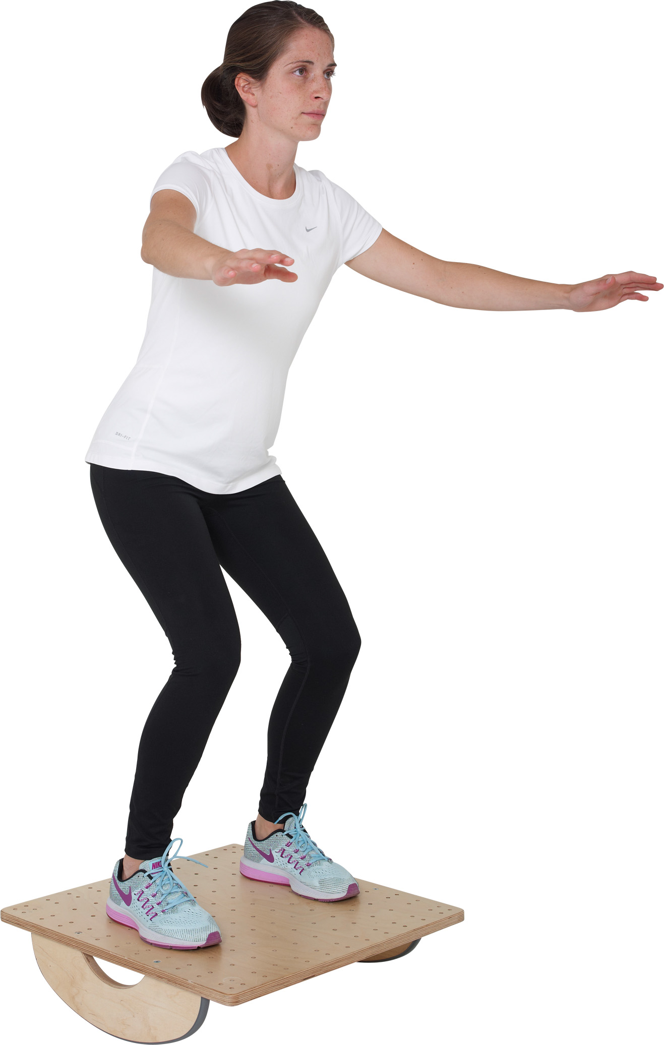A woman in sportswear is balancing on a light brown Pedalo PT wobble board. She is standing upright with her knees slightly bent and her arms outstretched. The board is square with holes and has two curved runners.