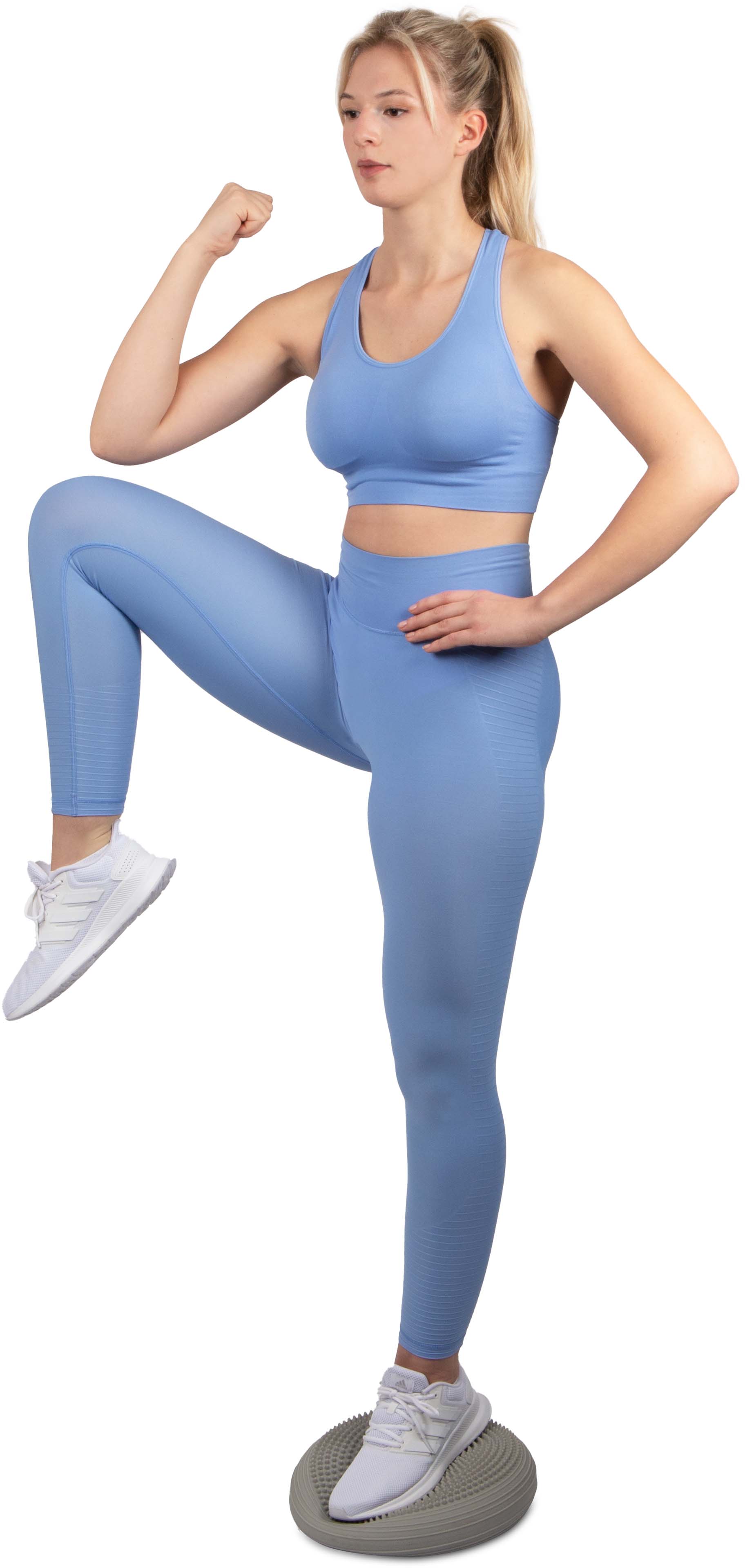 A blonde woman in light blue sportswear is standing on one leg on a grey Bamusta Coxim balance cushion, knee bent, fist raised, hand on her hip. An exercise for balance, stability and strength.