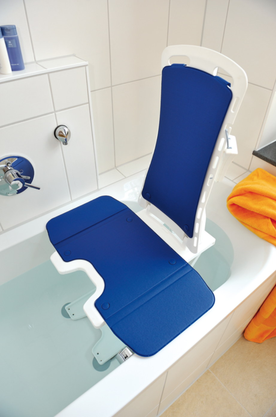 Sollevatore per vasca da bagno Bellavita blu in una vasca bianca piena d'acqua. Struttura bianca con seduta e schienale imbottiti di colore blu, con apertura igienica visibile. Per entrare e uscire in sicurezza. Piastrelle bianche, asciugamano arancione.