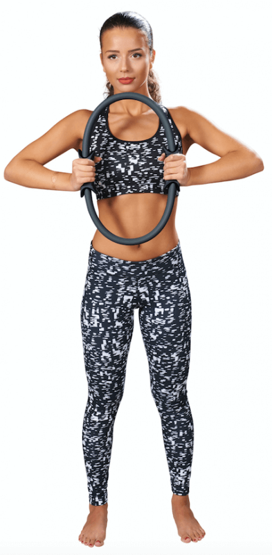 Une femme sportive en tenue de fitness noire et blanche tient un anneau de Pilates gris à hauteur de poitrine. Elle démontre un exercice pour renforcer les abdominaux et le dos. Anneau à poignées caoutchoutées.