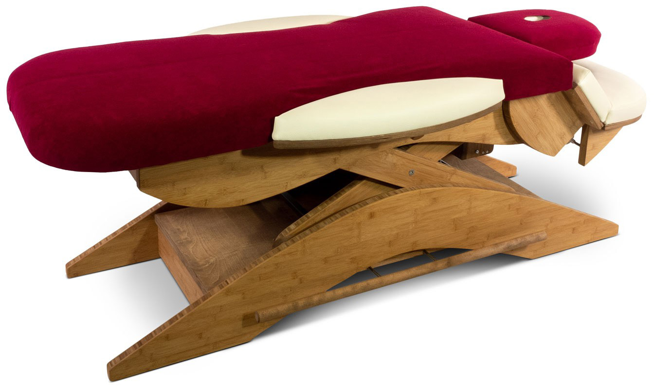 A modern treatment couch with a wine-red, velvety cover and cream-coloured accents. The frame is made of light brown, curved wood, revealing an adjustable mechanism. The couch is ready for therapy or relaxation.