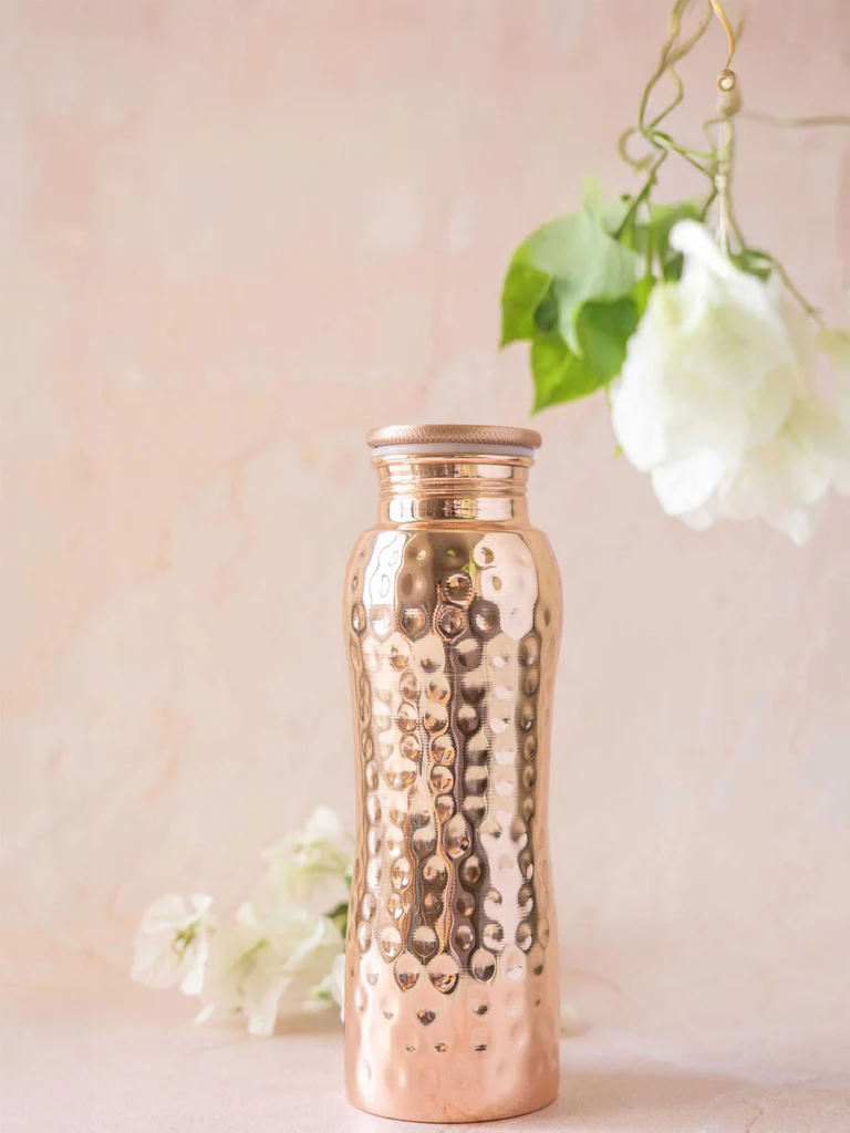 Glänzende, kupferfarbene Wasserflasche im Curve-Design mit markanter, handgehämmerter Oberfläche, die das Umgebungslicht lebendig reflektiert. Auf dezent hellrosa Untergrund, mit unscharfen weissen Blüten und grünen Blättern im Hintergrund.