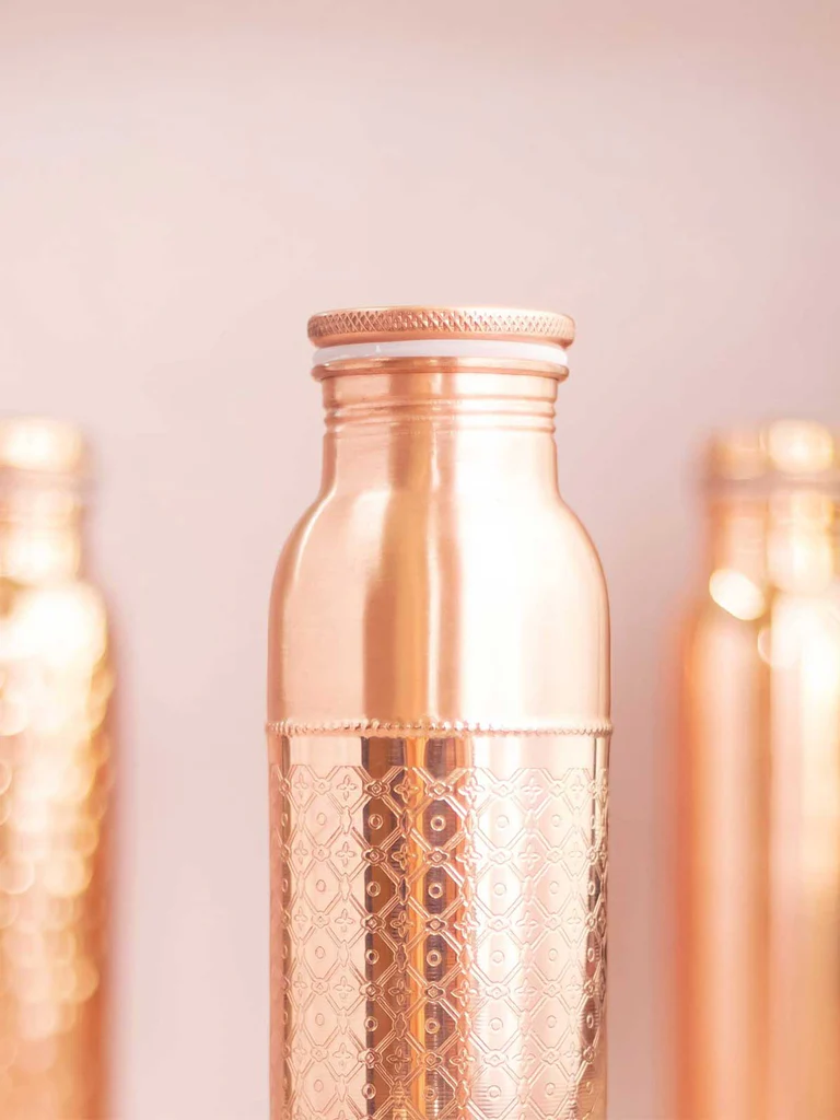 Glänzende roségoldene Kupferflasche mit feinem, handgeätztem Mosaikmuster. Schraubverschluss, Lichtreflexe. Heller rosa Hintergrund mit unscharfen Flaschen.