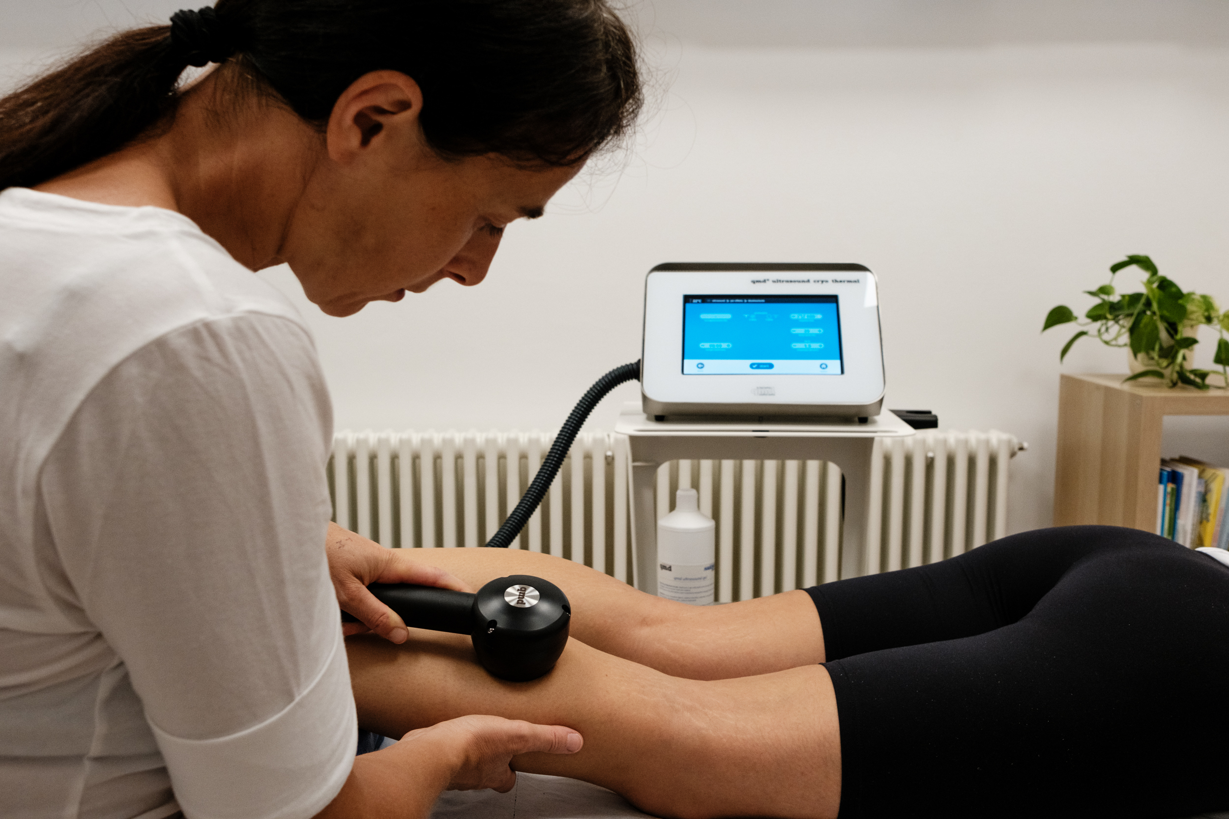 Eine Therapeutin behandelt das Bein einer bäuchlings liegenden Patientin mit einem qmd® cryo-thermal Komplett-System. Im Hintergrund ist das Bediengerät mit Touchscreen auf dem Gerätewagen zu sehen. Gel-Flasche und Heizkörper ebenfalls.