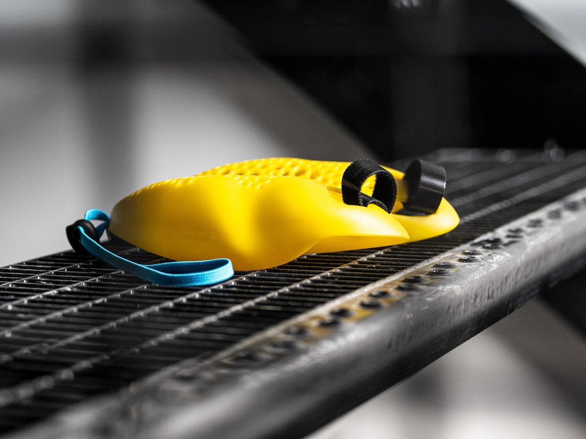 Deux poignées ergonomiques jaune clair reposent sur une grille métallique foncée. L’une avec dragonne bleue, l’autre avec dragonne noire pour fixation aux mains. Leur forme améliore posture et efficacité en marche ou course.