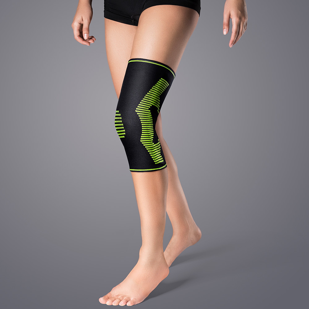 A person wears a black knee support on their right knee with light green diagonal stripes. DermaPlast Active Genu Soft plus stabilises and relieves knee joint pressure, offering targeted compression, a cool fit and silicone strips.