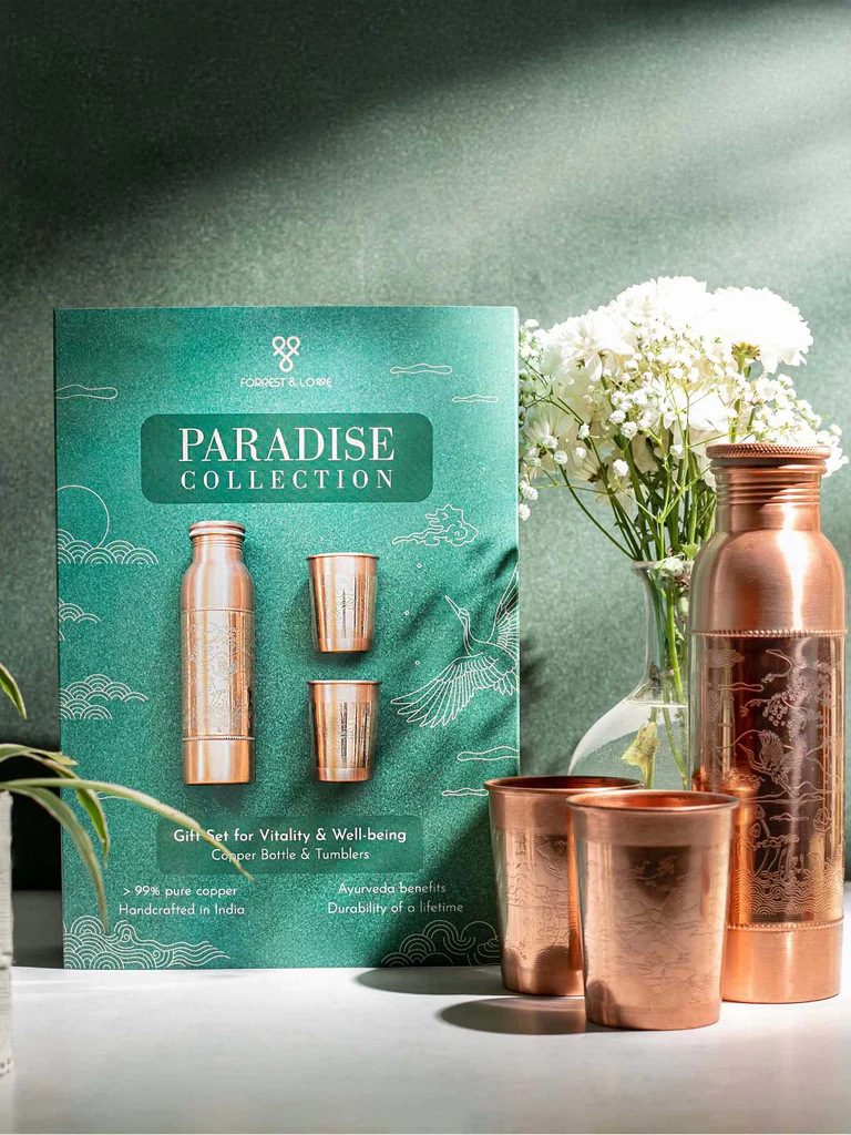 Geschenkset mit kupferner Flasche und zwei Bechern im eleganten Paradies-Design vor passender grüner Verpackung. Flasche und Becher mit fein geätzten Vogel- und Blumenmustern. Rechts im Hintergrund: Strauss weisser Blüten in Glasvase.
