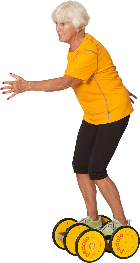 An elderly white-haired woman in a yellow sports top and black capri trousers balances on a Pedalo Classic trainer. Feet on wooden footplates with yellow rollers, arms out for balance, leaning slightly forwards.