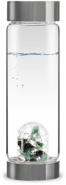 A tall, transparent VitaJuwel ViA glass water bottle, about two-thirds full of clear water. At the bottom, a cylindrical glass insert containing greenish and light-coloured stones. Small air bubbles rise to the surface. Grey metal cap and base.