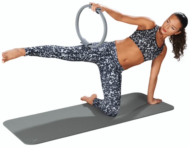 Femme sportive fait un exercice de Pilates avec un anneau gris sur un tapis gris. Jambe droite tendue et levée vers l’arrière, anneau maintenu contre la cuisse droite par le bras gauche. Renforce le tronc, utile en rééducation.