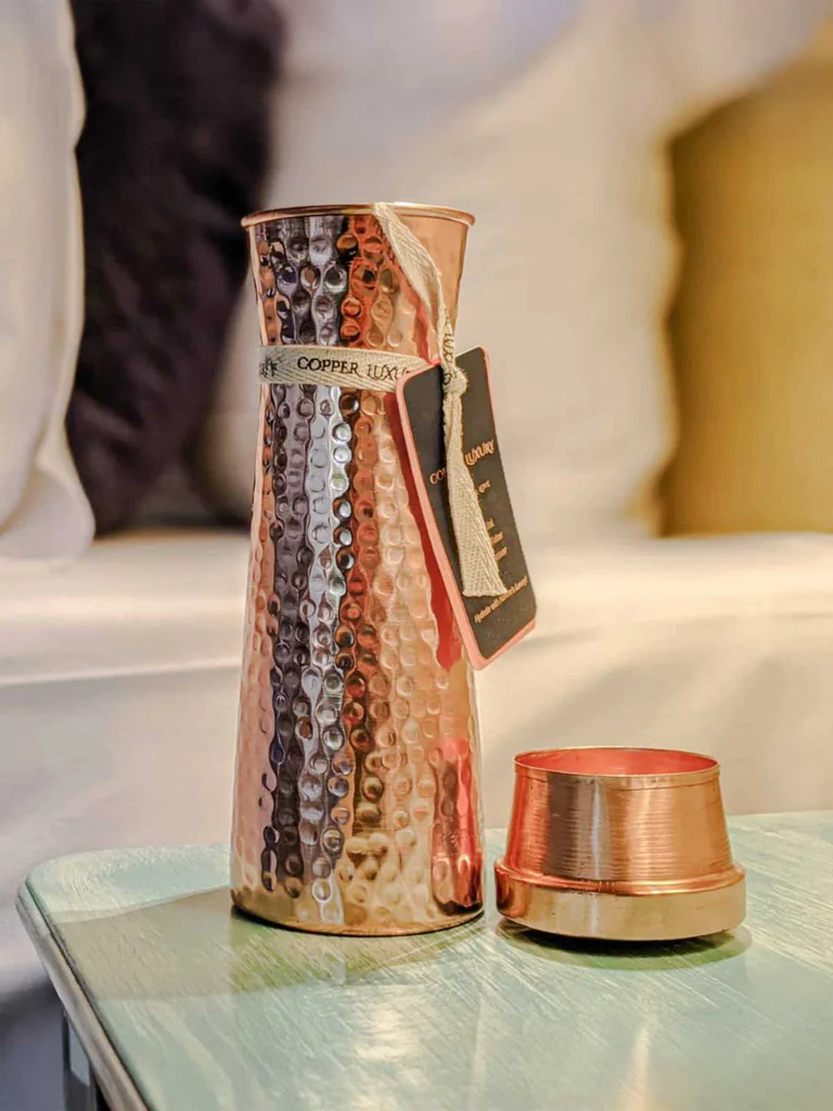Hochglänzende, bauchige Kupferkaraffe mit gehämmerter Oberfläche auf hellblauem Holztisch, daneben kleiner Kupferbecher. Um den Hals cremefarbenes Band mit „Copper Luxury“ und dunkelroter Anhänger. Hintergrund unscharf, helle Stoffe.