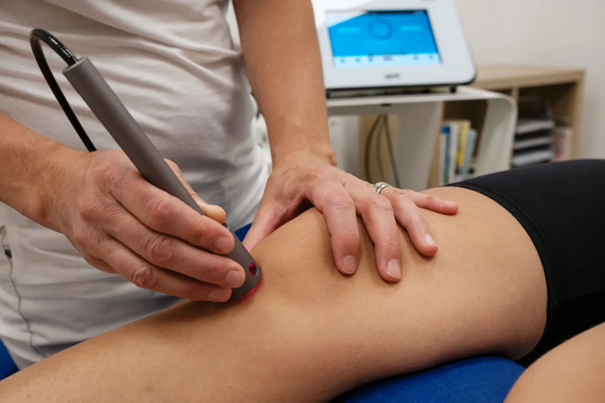 Eine Therapeutin nutzt ein qmd® helios Lasertherapie-Gerät am Oberschenkel eines Patienten. Die hell rot leuchtende Spitze berührt die Haut, während sie es präzise führt. Im Hintergrund ist der Bedienbildschirm zu sehen.