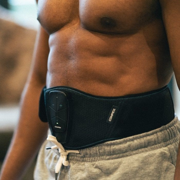 A muscular black man is wearing a black Compex CoreBelt 5.0 muscle stimulation belt around his waist. The belt has a control unit with buttons. He is wearing grey jogging bottoms and is bare-chested.