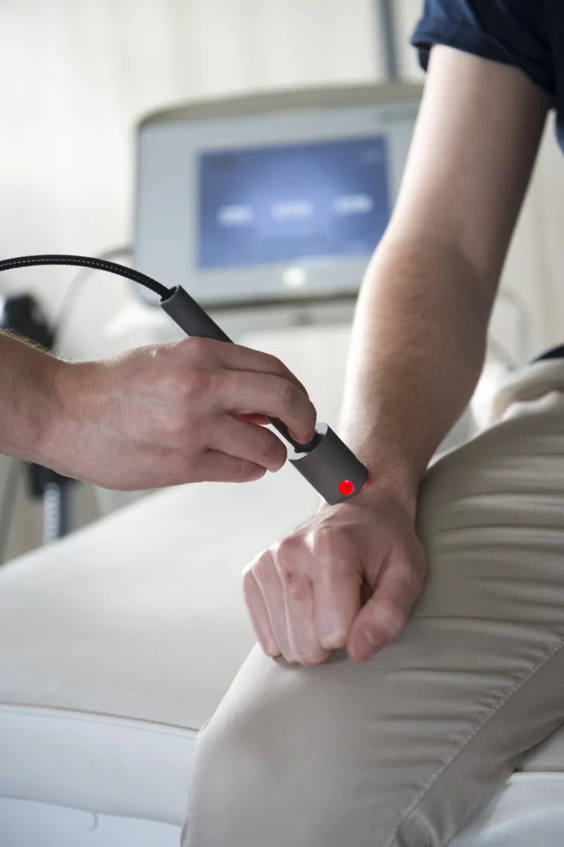 Helle Nahaufnahme: Eine Hand hält ein graues qmd® helios Lasertherapie-Gerät auf das Handgelenk eines Patienten. Ein roter Lichtpunkt markiert den Hautkontakt und deutet auf eine Therapie hin. Unscharf: medizinischer Bildschirm.