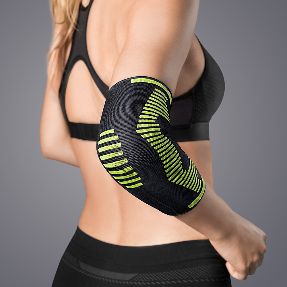 A woman is wearing a black elbow support with neon-green stripes on her right arm, which wraps around her bent elbow. The knitted material provides support and is designed for therapy or sport, with a focus on comfort and fit.
