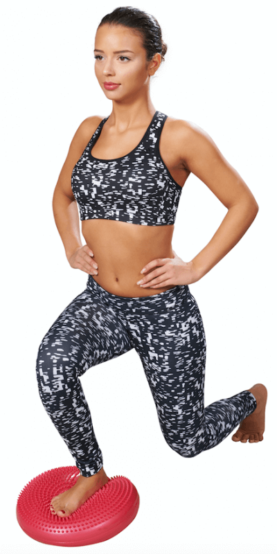 A woman dressed in sportswear – leggings and a sports bra – is performing a lunge. Her left foot is bare on a round, red balance cushion with nubs. With her hands on her hips, she is practising coordination, balance and strengthening her muscles.