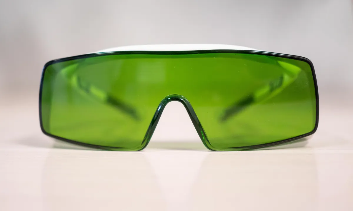 Frontansicht einer rechteckigen Laserschutzbrille mit dunkelgrün getönten, breiten Gläsern und einem weissen Oberrand, die auf einer hellen Fläche liegt. Sie ist speziell für den Augenschutz während Lasertherapie-Anwendungen konzipiert.