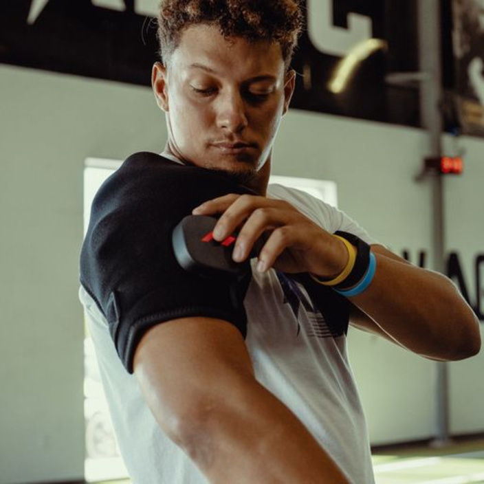A man is using a Hyperice Venom 2 shoulder massage belt on his left shoulder and is pressing a small, dark control unit against the black belt. He is wearing a white T-shirt and coloured wristbands, in a gym setting.