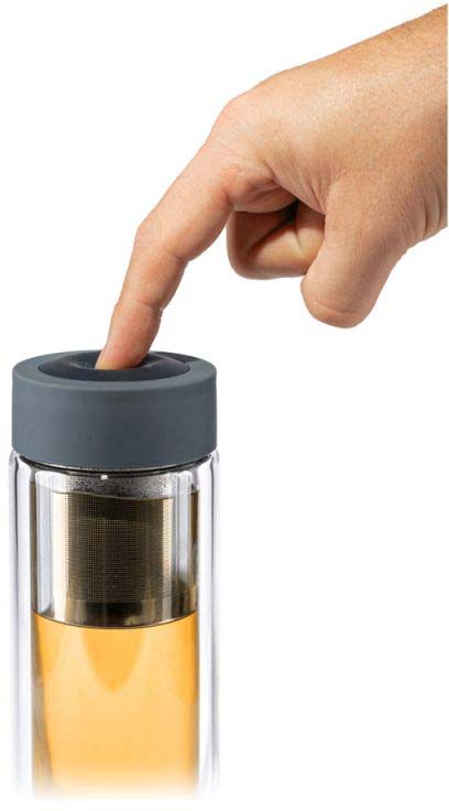 Una mano preme sul coperchio scuro in silicone di una borraccia trasparente in vetro a doppia parete. All’interno si vede un sottile colino da tè in acciaio inossidabile con tè sfuso immerso in un liquido color oro-marrone. Facile da usare.