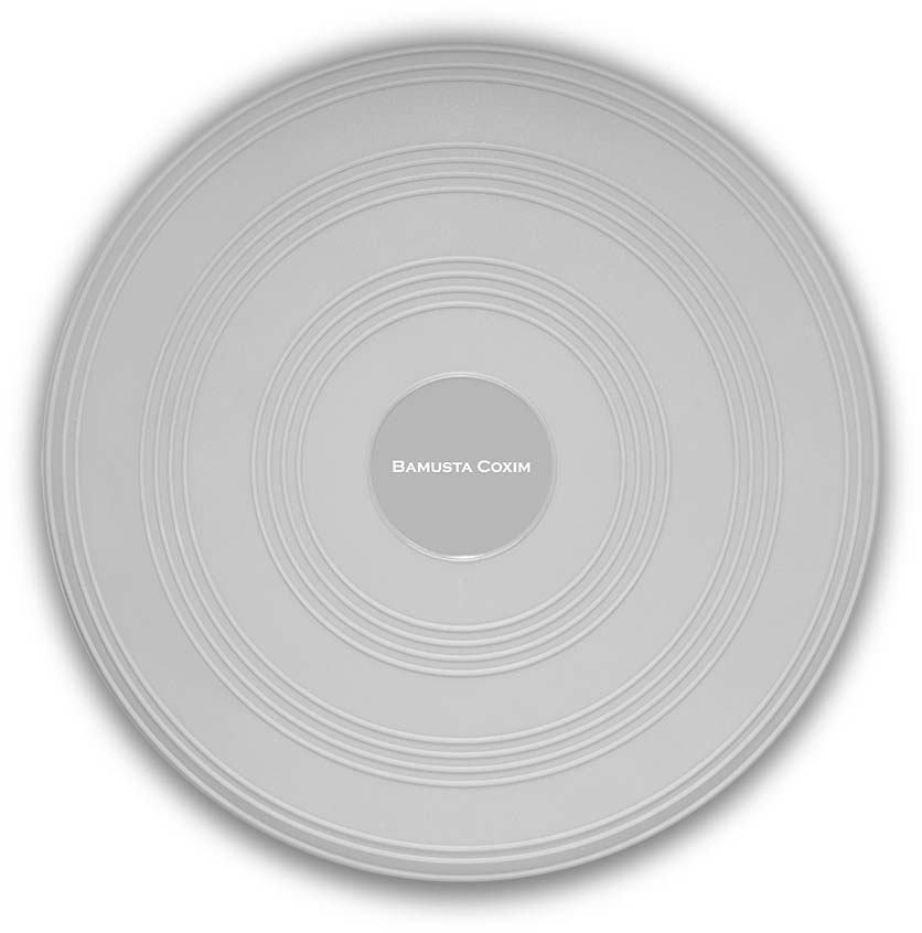 A light grey, round Bamusta Coxim balance cushion lies flat against a light-coloured background. The surface features concentric, raised grooves. In the centre is a smooth, slightly darker area bearing the words ‘BAMUSTA COXIM’ in white.