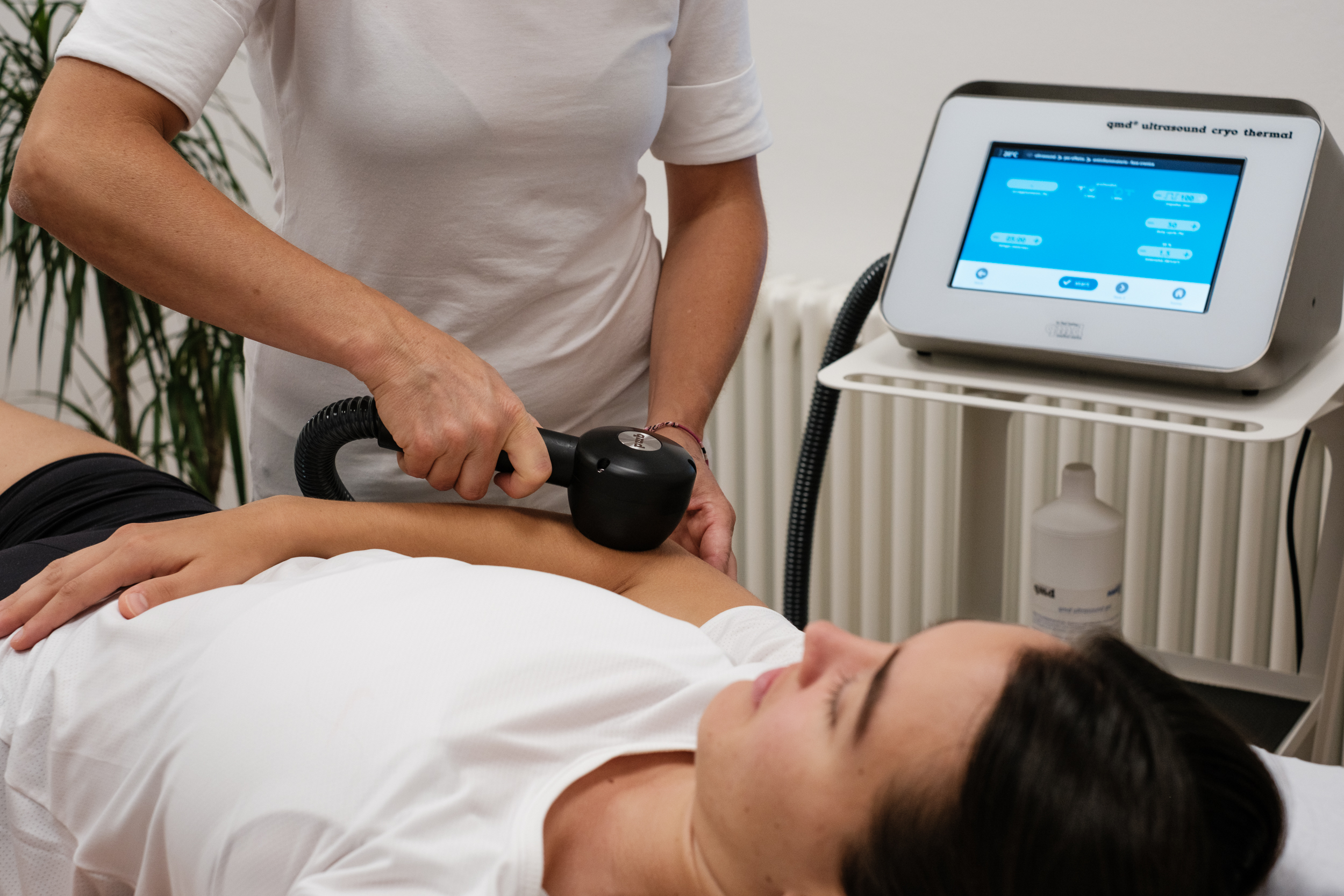 Therapeutin in weisser Kleidung behandelt den Oberarm einer Patientin auf der Liege mit dem schwarzen Handstück des qmd® cryo-thermal Komplett-Systems. Im Hintergrund: Steuergerät mit Display, Gerätewagen und Ultraschallgel.