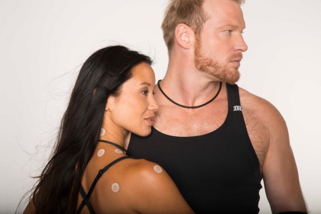 Close-up of a couple wearing Phiten Titan Ban Power Tapes. The woman, with long dark hair, has round, skin-coloured tapes on her neck and shoulders. The bearded man is wearing a tape around his neck. Both are dressed in black and look relaxed.