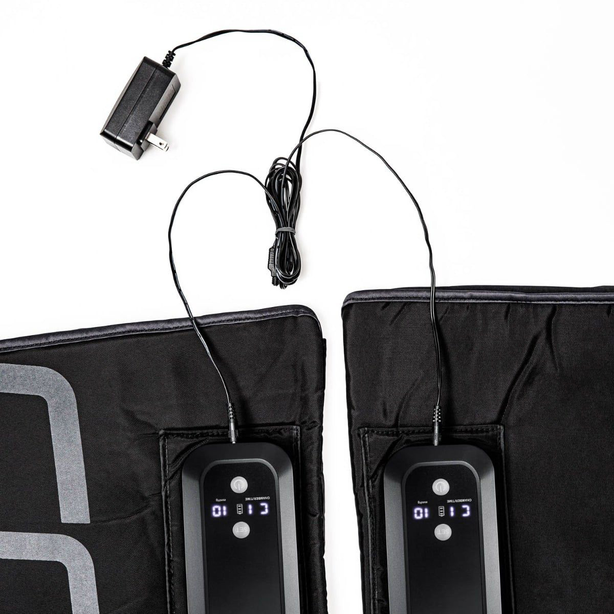 Two black Compex Ayre compression boots on a white background. Each cuff has an integrated control unit with a digital display (‘C 1 B 10’, ‘mmHg’). The charging cable and mains adaptor are shown above. Wireless compression for recovery.