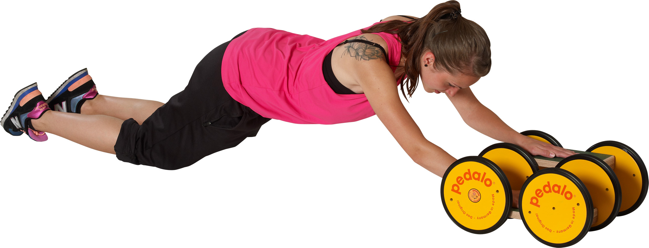 A young woman in a pink sports top and black shorts is doing a full-body exercise: lying face down with arms stretched out, she pushes the yellow Pedalo Classic with wooden base and twin wheels forward to improve balance and core strength.