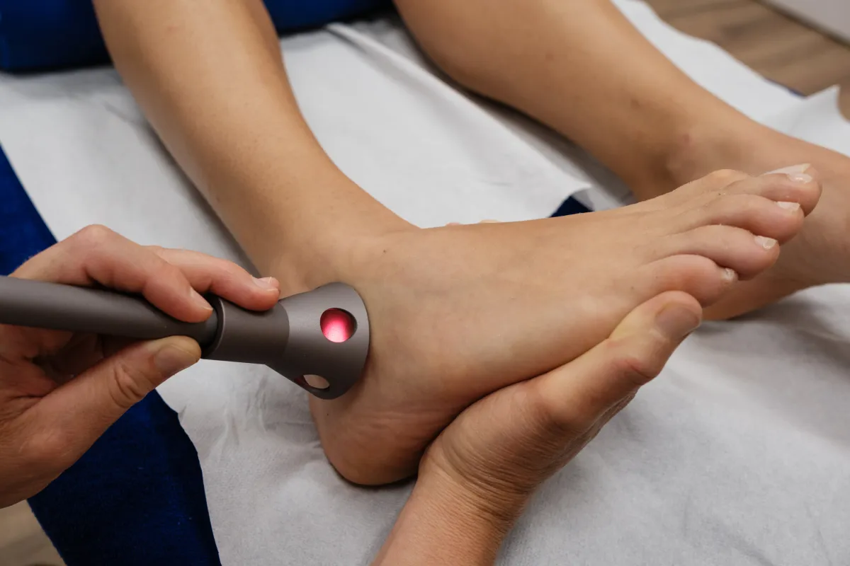 Eine Person erhält eine qmd Lasertherapie am Sprunggelenk des rechten Fusses. Eine Hand hält ein graues, schlankes Gerät mit hellrot leuchtendem Applikator, präzise auf die Haut gerichtet. Der Fuss liegt entspannt.