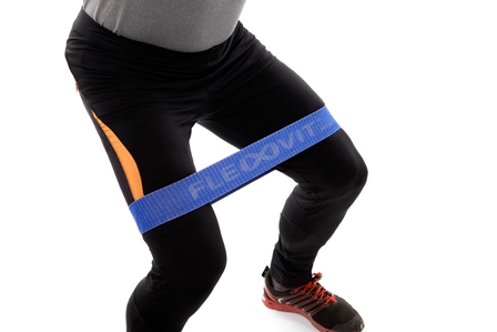 Une personne en tenue de sport, vêtue d'un pantalon noir, utilise une bande de résistance bleue FLEXVIT Mini autour des cuisses, au-dessus des genoux. La posture suggère un exercice visant à renforcer et à stabiliser les muscles des jambes et des hanches.