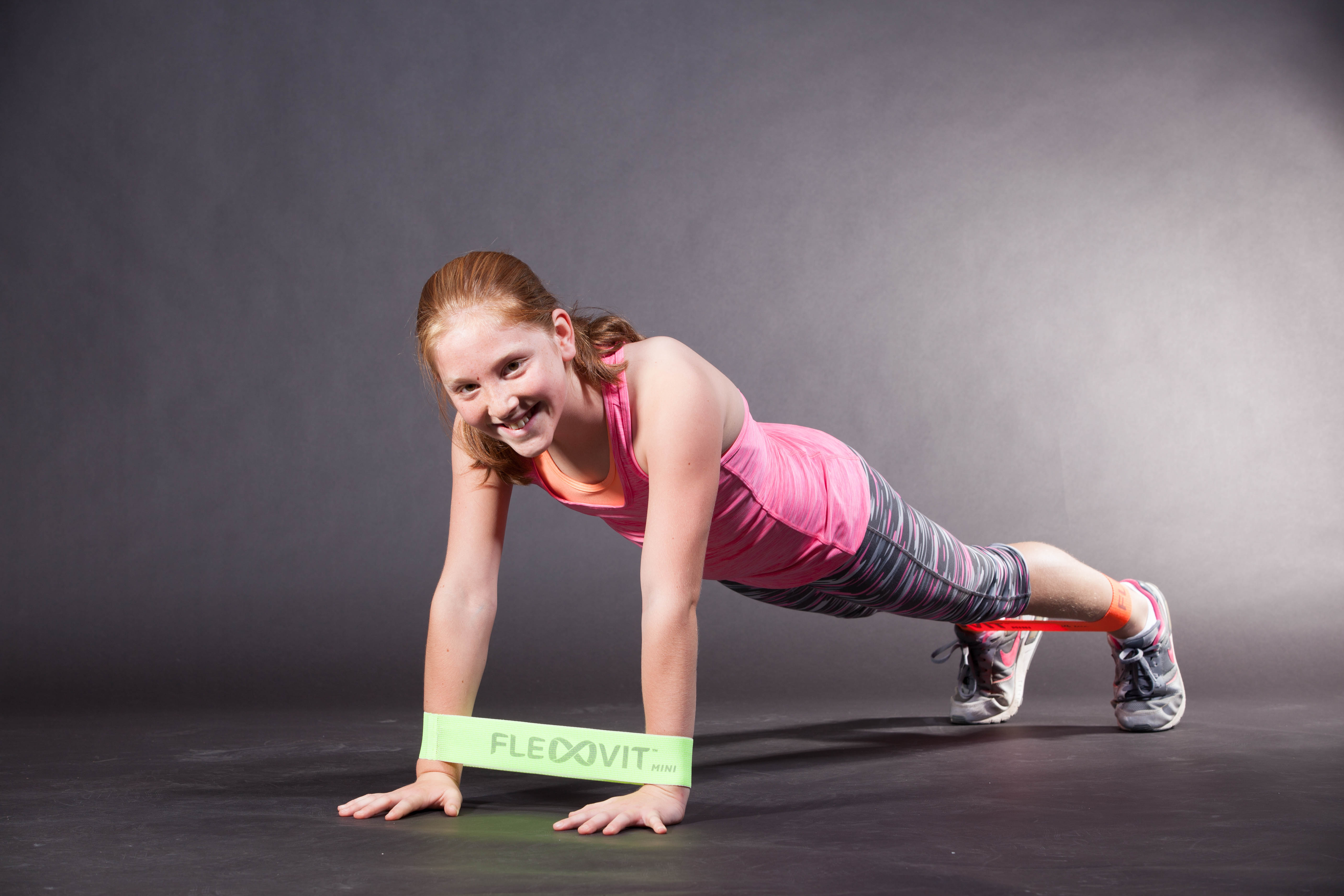 Une jeune personne souriante fait une planche. Une bande FLEXVIT Mini vert clair est enroulée autour des poignets, et une bande rouge autour des chevilles. La personne porte une tenue de sport et s'équilibre sur un sol sombre.
