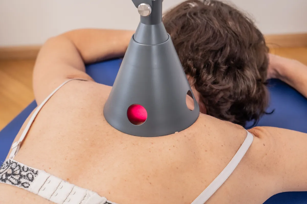 Eine Person liegt auf dem Bauch auf einer blauen Behandlungsliege. Ein graues, konisches Lasertherapiegerät strahlt ein rotes Licht auf den oberen Rücken der Person. Das Gerät ist so positioniert, dass der Laser direkt auf die Haut der Patientin wirkt.
