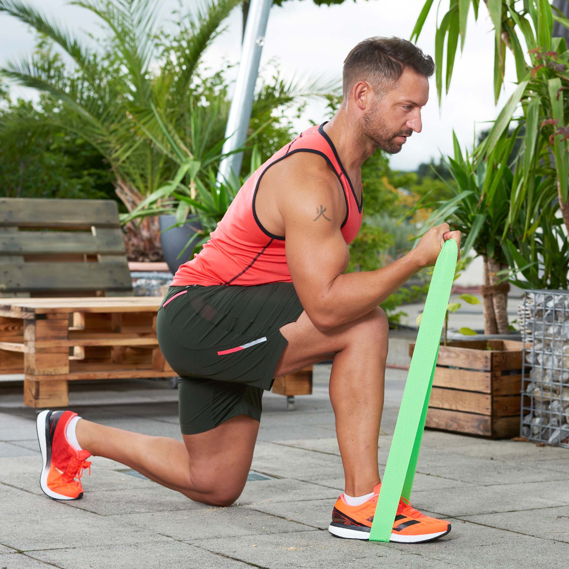 A man is kneeling outdoors and exercising with a green fitness band: the band is secured under his foot and he is pulling it towards his chest with both hands. Plants and wooden pallets can be seen in the background.