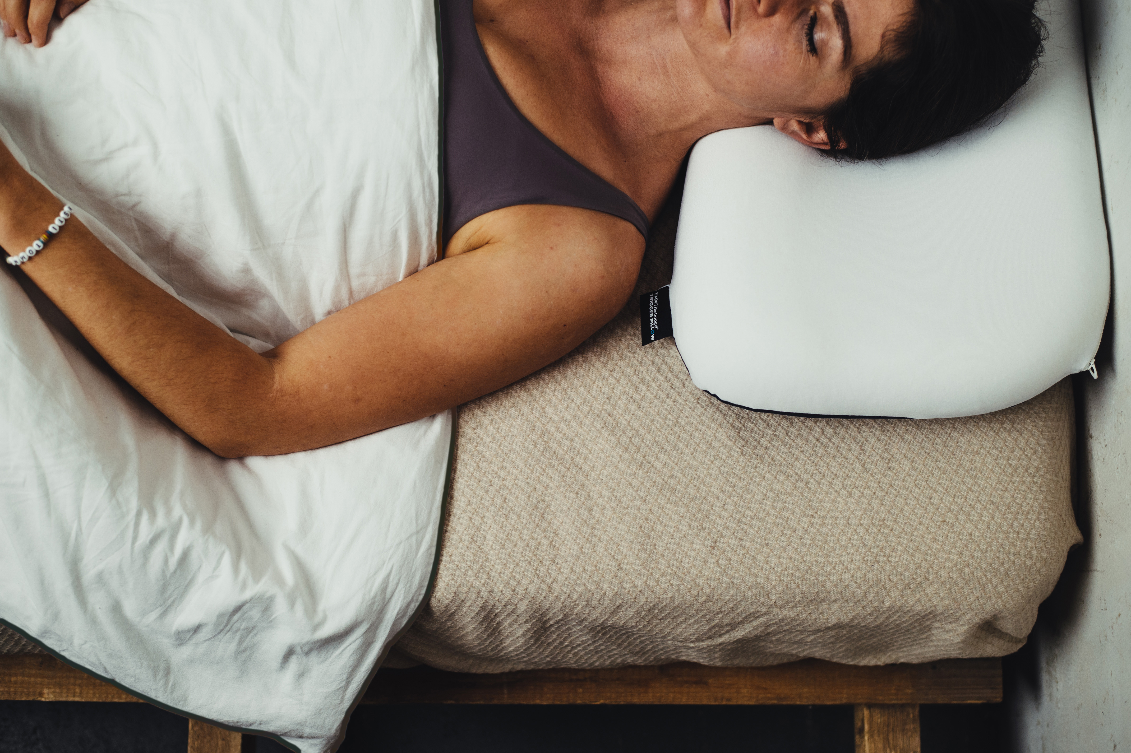 A person is sleeping peacefully on their side in bed, covered with a white, crease-resistant TMX® Thermo Cover duvet. Their head rests on an ergonomic pillow. The duvet covers their upper body and one arm; a beige mattress topper is visible underneath.