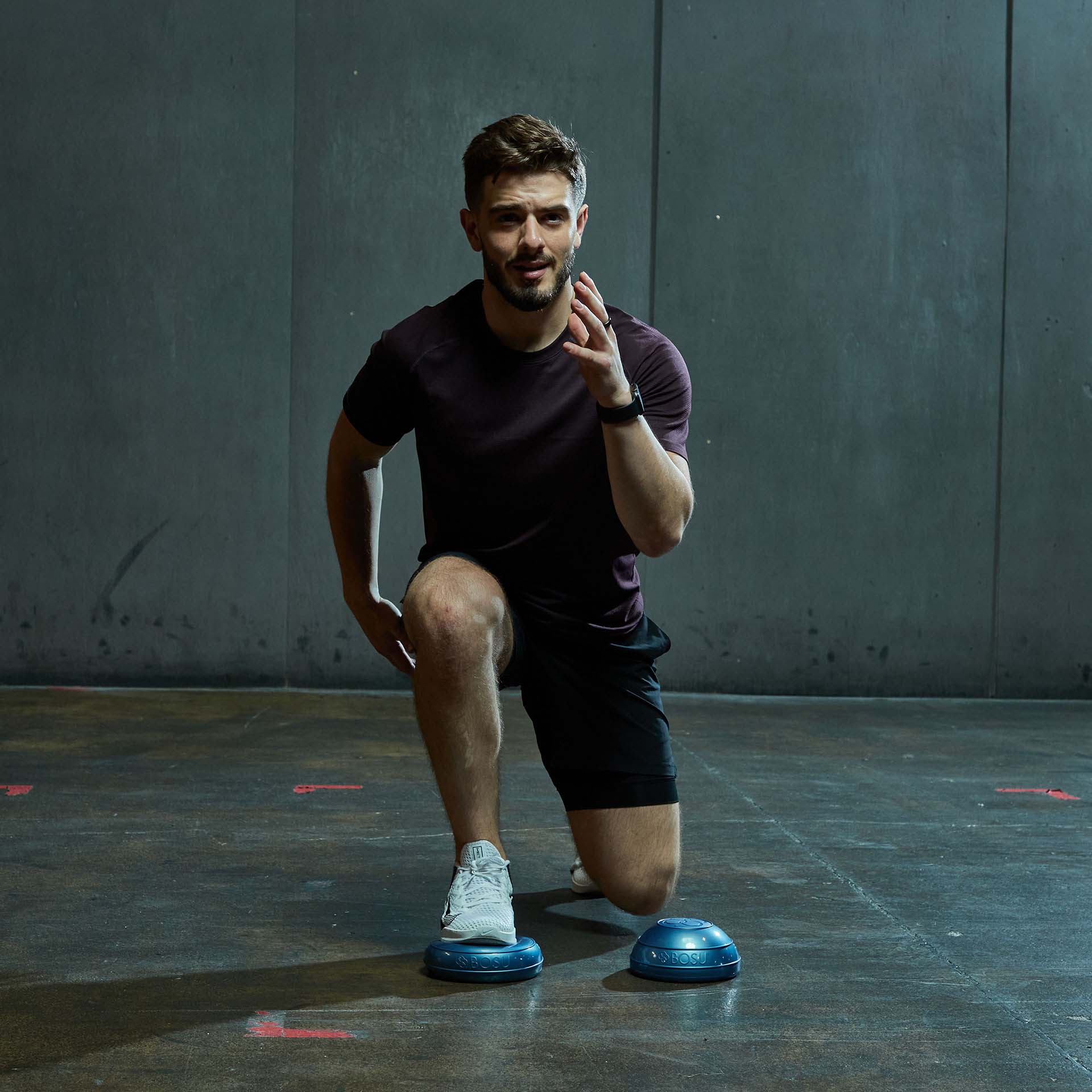 A sporty man is kneeling in a lunge position: his left foot is on a blue Balance Pod, with a second Pod on the floor next to it. He is looking straight ahead and practising his balance, coordination and stability in front of a dark wall.